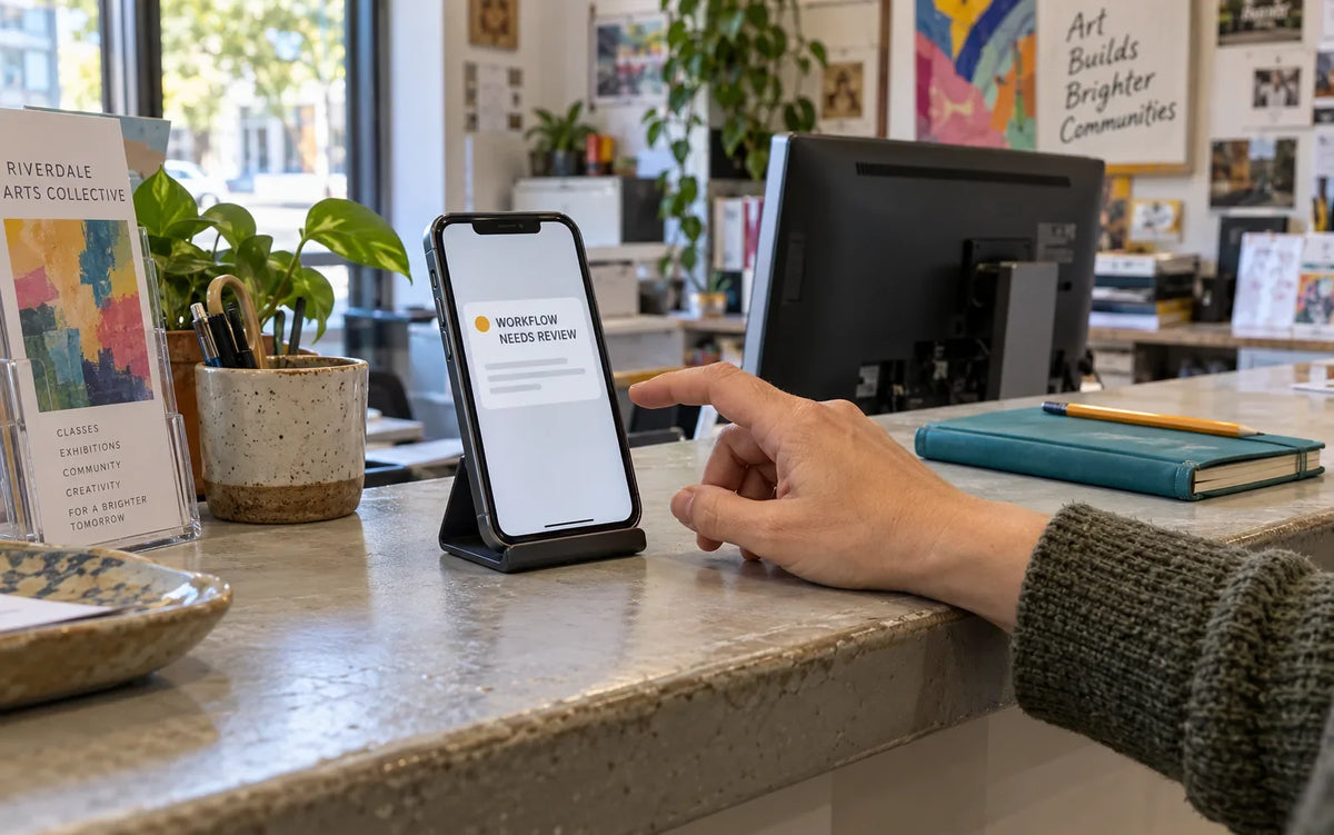 A hand beside a smartphone displaying an illustrative workflow-review notification at an arts office reception counter.