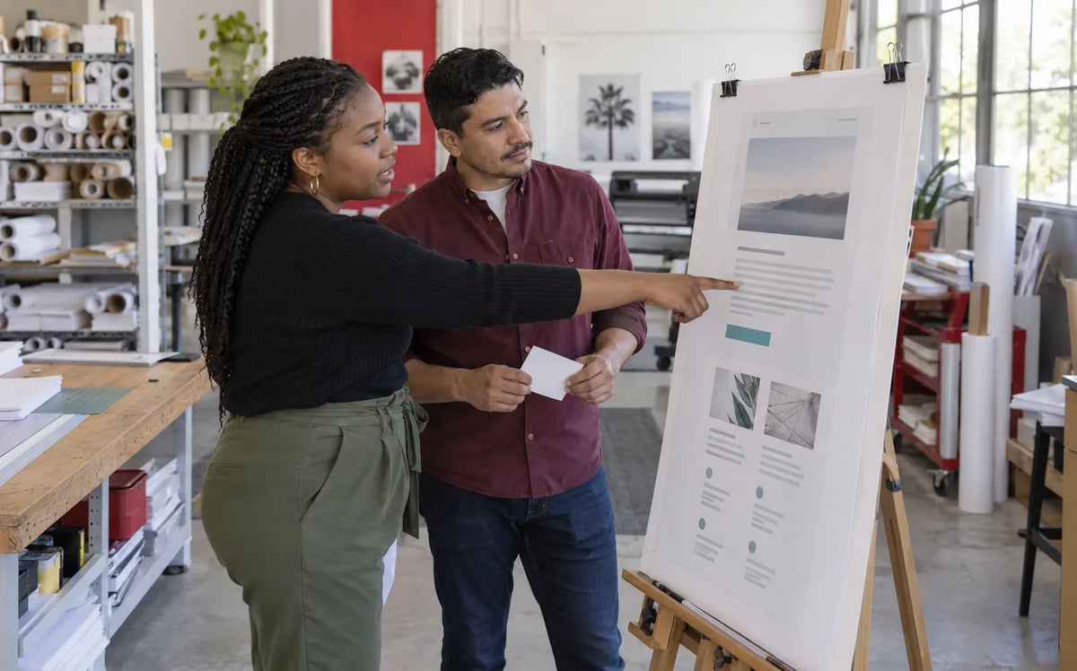 Two colleagues review a printed page layout on an easel in a print workshop